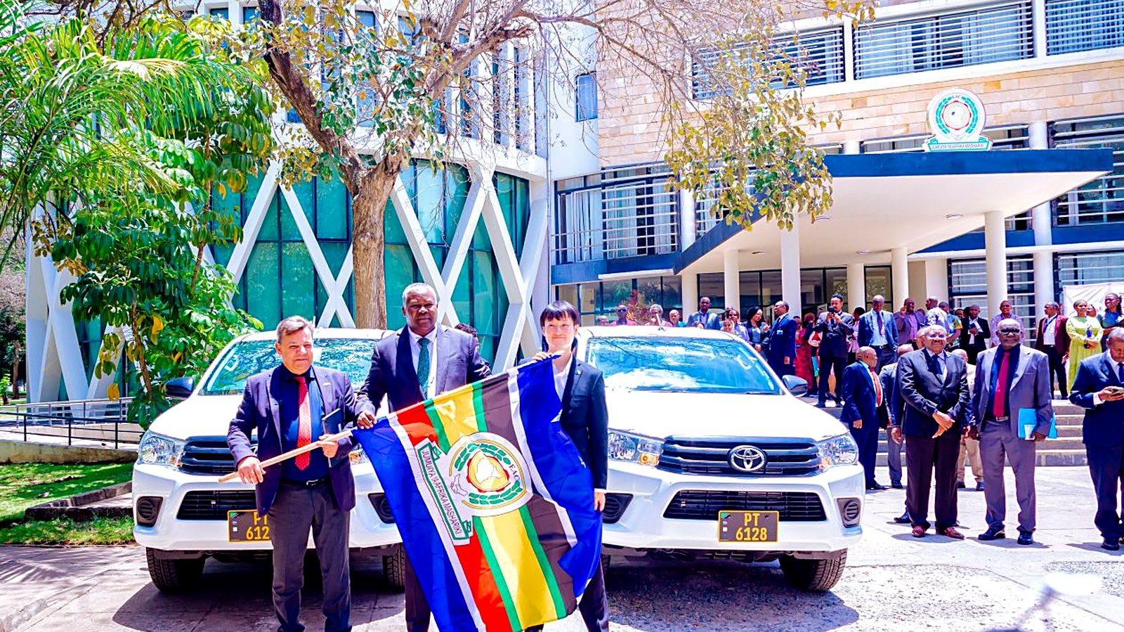 Officials hold the East African Community flag in front of two of the 16 Japan-funded patrol vehicles at the EAC headquarters in Arusha, Tanzania. Each of the eight Partner States is receiving two vehicles for use at border points.